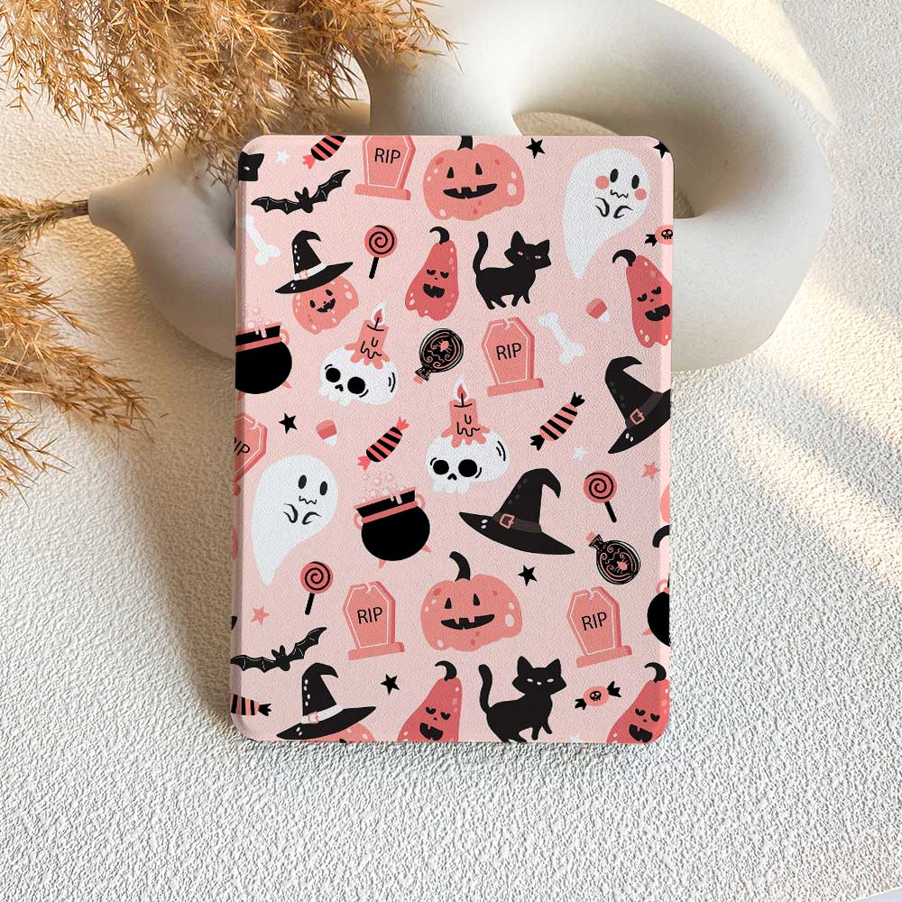 A pink Kindle case with a pattern of Halloween-themed characters and motifs, including ghosts, pumpkins, and cats, displayed on a table with a decorative background.