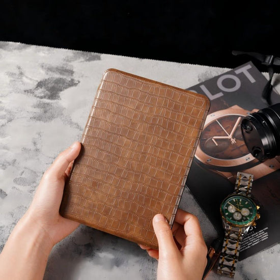 Brown crocodile-patterned wallet held by hands on a marble surface with a watch and camera in the background.