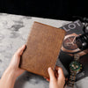 Brown crocodile-patterned wallet held by hands on a marble surface with a watch and camera in the background.