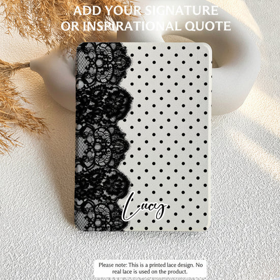 A tablet case with a black lace panel on the left side and a white background with black polka dots on the right, shown on a textured light surface with decorative dried grass and a white ceramic object. Text at the top says 'ADD YOUR SIGNATURE OR INSPIRATIONAL QUOTE' and the name 'Lucy's' is printed on the case.