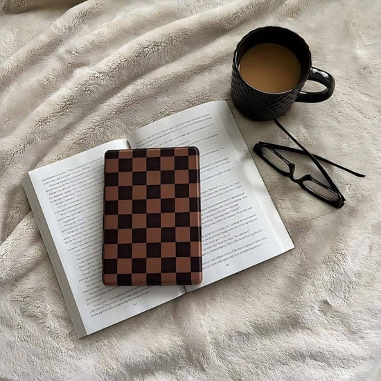 Open book with a checkered kindle case object on top, next to a black mug and glasses on a soft surface.