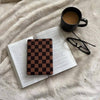 Open book with a checkered kindle case object on top, next to a black mug and glasses on a soft surface.