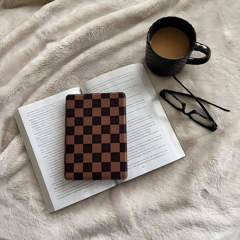 Open book with a checkered kindle case object on top, next to a black mug and glasses on a soft surface.